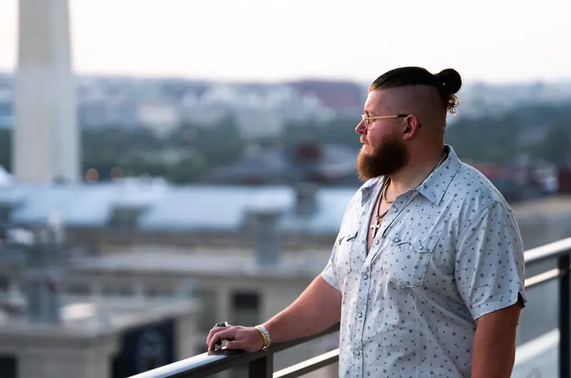 Luke leaning on a rooftop railing above a city skyline, looking off to one side.
