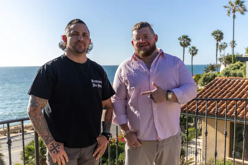 Two men standing together on a coastal terrace, one gesturing toward the other, ocean and palms behind them.
